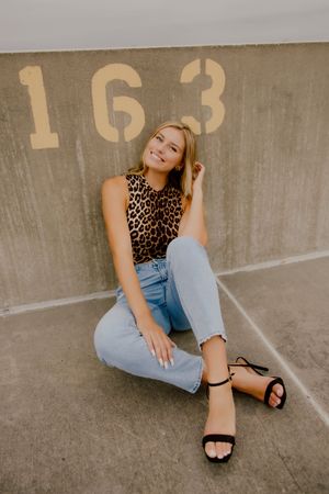 Blonde teen in leopard print top and jeans sits against concrete wall with number 163.