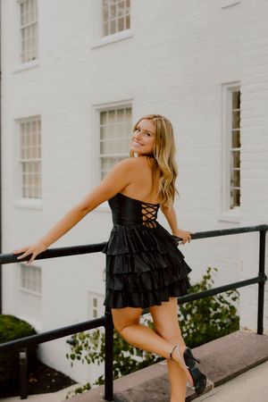 Young woman in black strapless ruffled dress posing by iron railing outside white brick building.