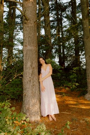 Brunette woman in pink floral dress leaning against tall tree trunk in serene forest setting.