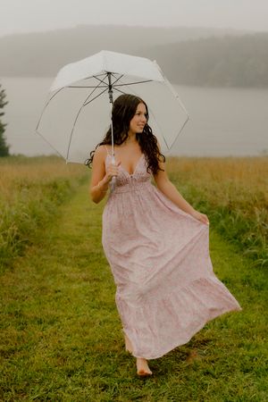 Brunette woman in pink floral maxi dress holding white umbrella walking through misty lakeside field.