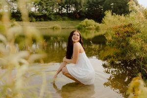 Young woman in white dress crouches smiling in shallow pond surrounded by lush greenery.
