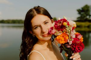 Brunette woman holds vibrant bouquet of colorful flowers near serene lake at golden hour.