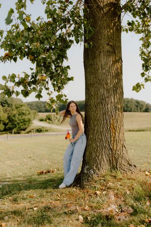 Young woman in gray top and jeans leaning against large tree trunk in sunny green field.