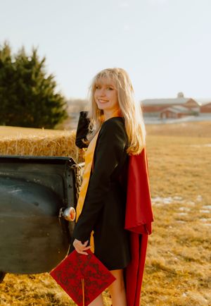 Smiling blonde graduate in black gown with red cape holding cap beside hay bale in golden field.
