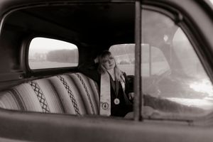 Black and white photo of blonde girl in graduation attire sitting in vintage car interior.