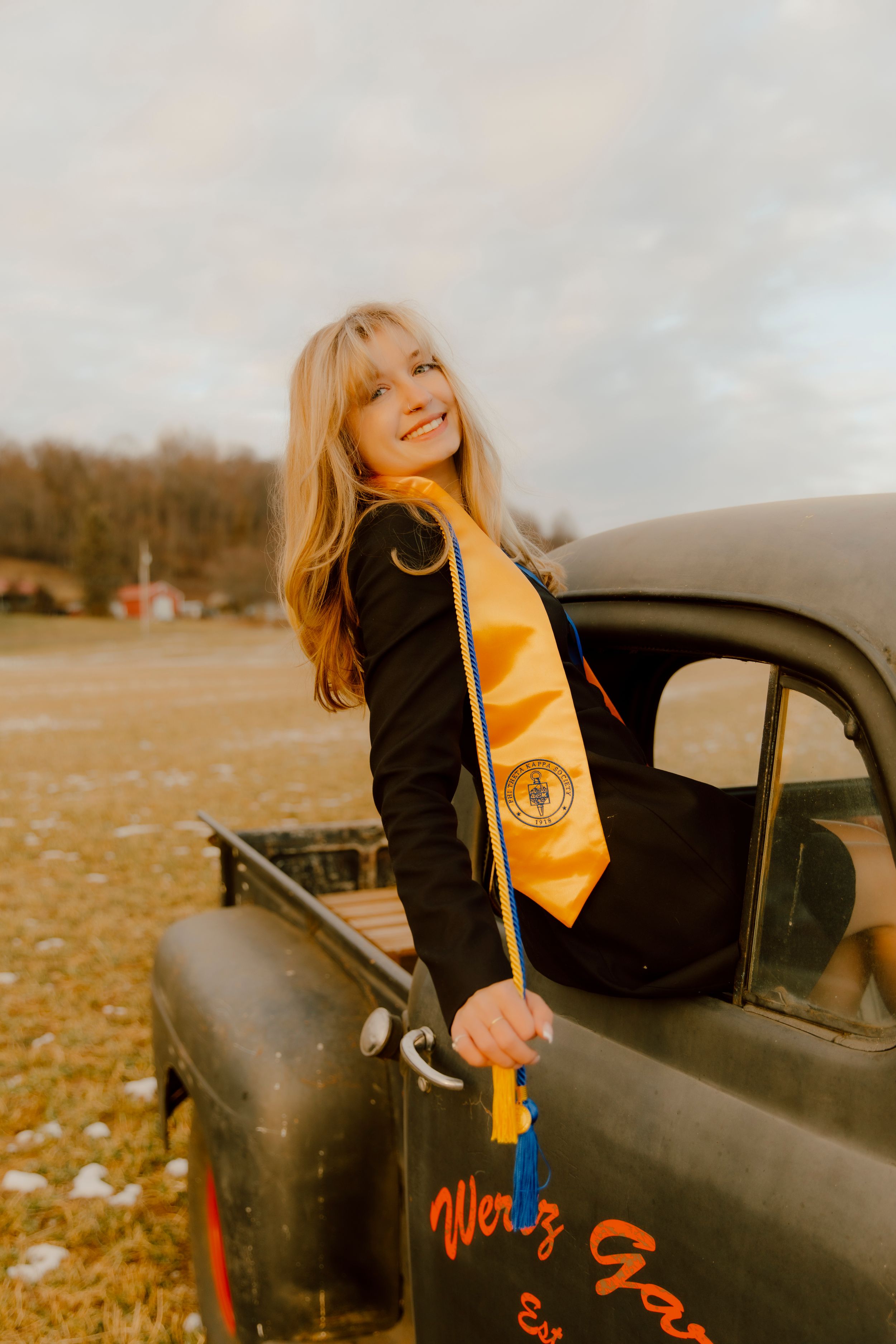 Smiling blonde graduate wearing gold stole leans out of vintage black truck in field.