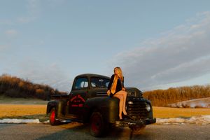 Young woman in black outfit sitting on vintage dark green truck at golden hour in rural landscape.