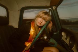 Young blonde woman rests against car seatbelt inside vehicle at golden hour sunset.