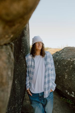 Curly-haired teen in plaid shirt, white tee, jeans and beanie leaning between large boulders outdoors.