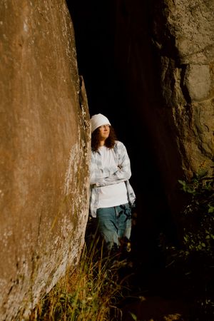 Young woman in white hat and casual clothes leaning against large rock formation in natural setting.
