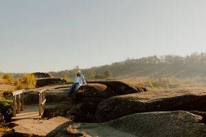 Person sitting on rocky outcrop overlooking misty scenic landscape with trees on clear morning.