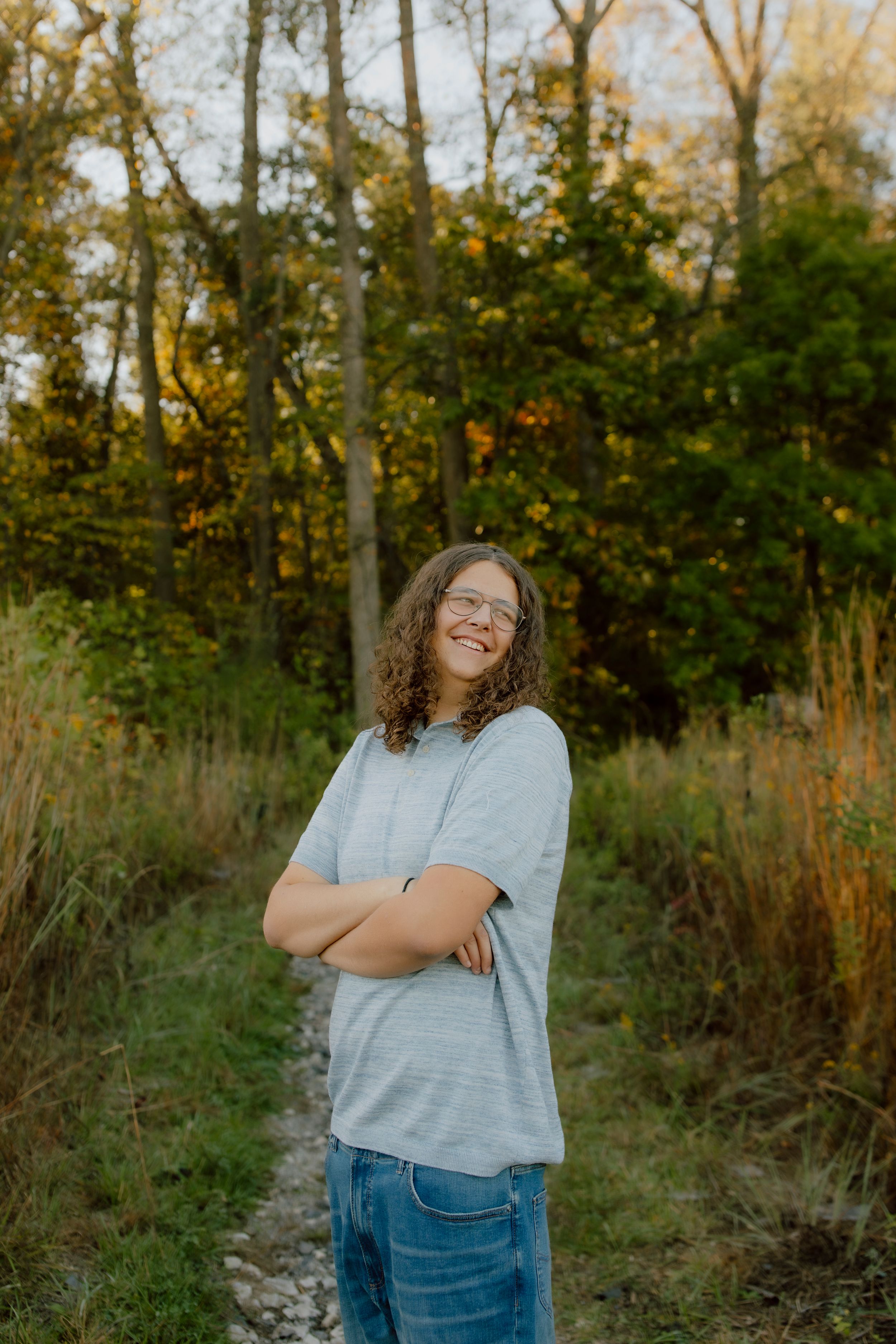 Curly-haired teen in gray shirt stands arms crossed smiling on woodland trail at sunset.