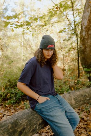 Young man with curly hair and glasses leans on log in autumn forest wearing beanie.