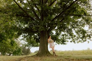 Young woman in lace dress leans against massive oak tree in open green field.