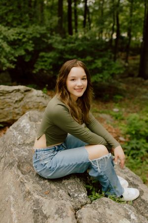 Smiling teenage girl in green top and ripped jeans sitting on rocks in a lush forest setting.
