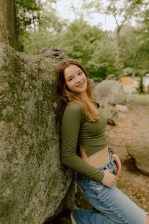 Teen girl in green long sleeve crop top and jeans leans against mossy boulder in forest.