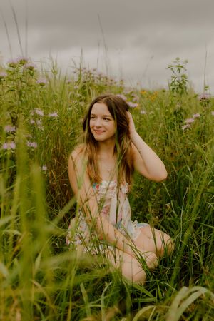 Teen girl in floral dress sits smiling in tall wildflower meadow under cloudy sky.