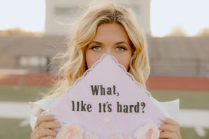 Blonde woman holds decorated graduation cap reading What like its hard outdoors at stadium.