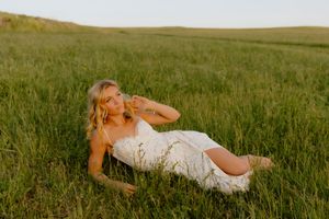 Young blonde woman in white lace dress reclines in lush green grass field at sunset.