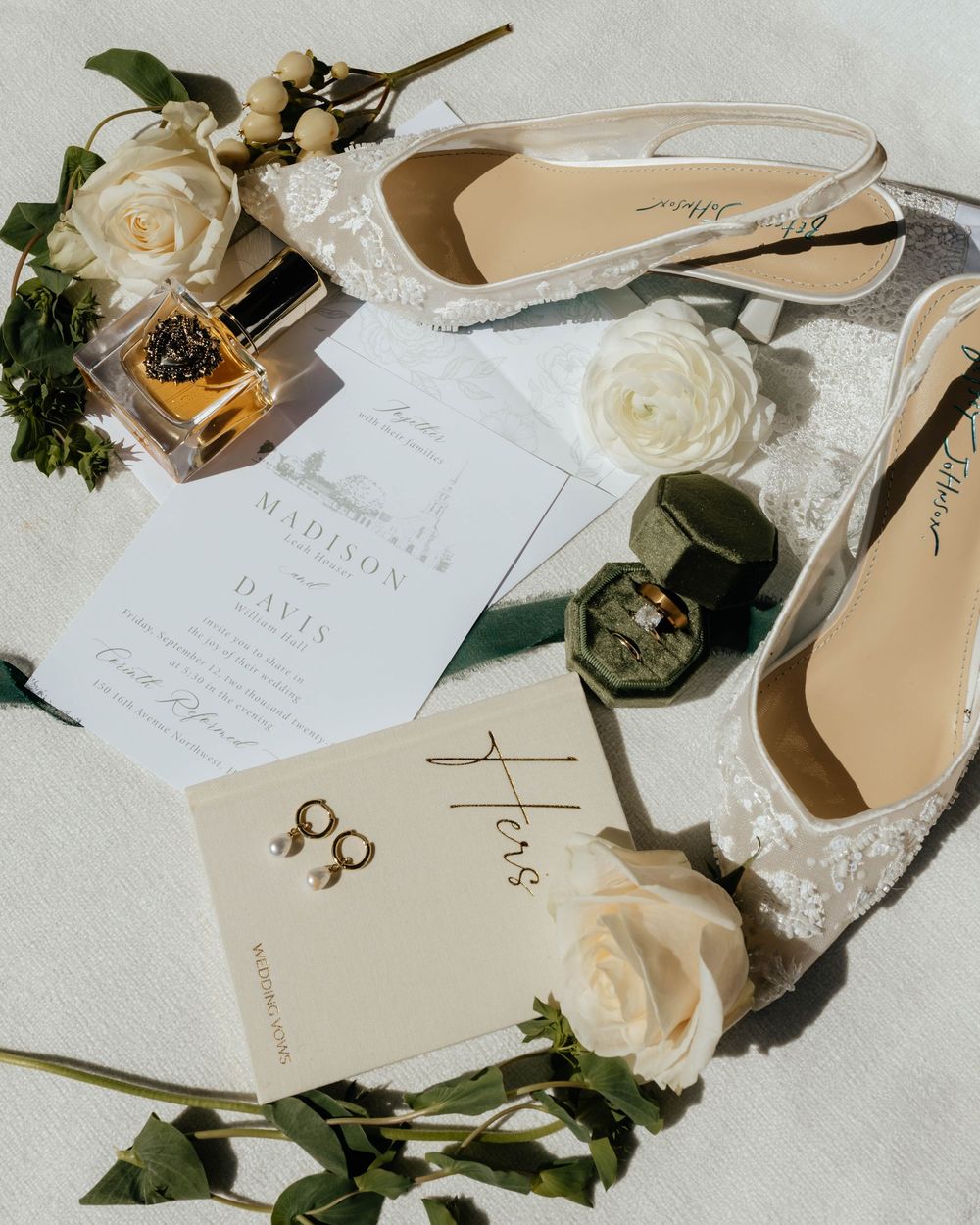 Wedding invitation suite with white roses, elegant calligraphy, and beige bridal shoes arranged on light surface.