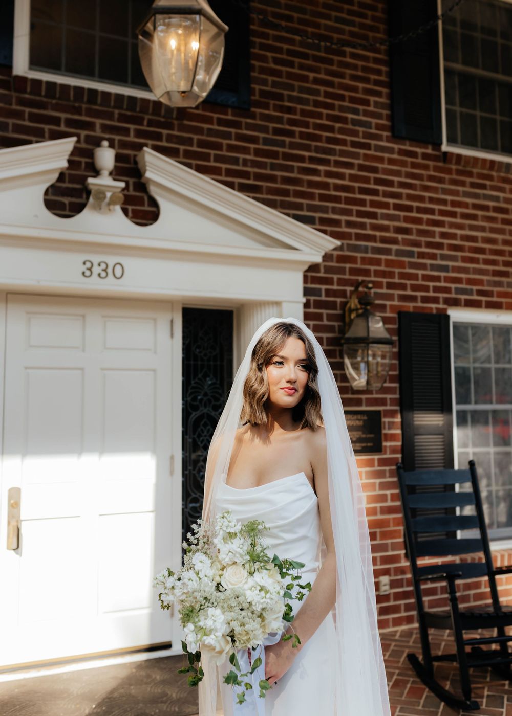 Elegant bride in white strapless gown holds white floral bouquet outside historic brick home with address number 330.