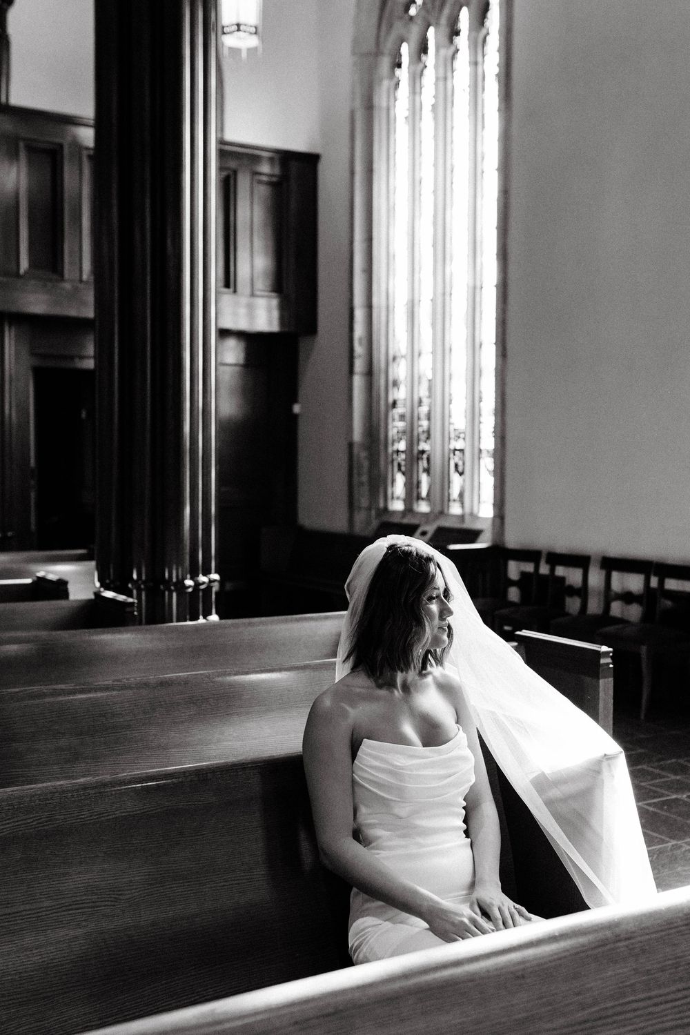 A bride in a white dress sits alone in a church pew, with tall stained glass windows visible in the background.