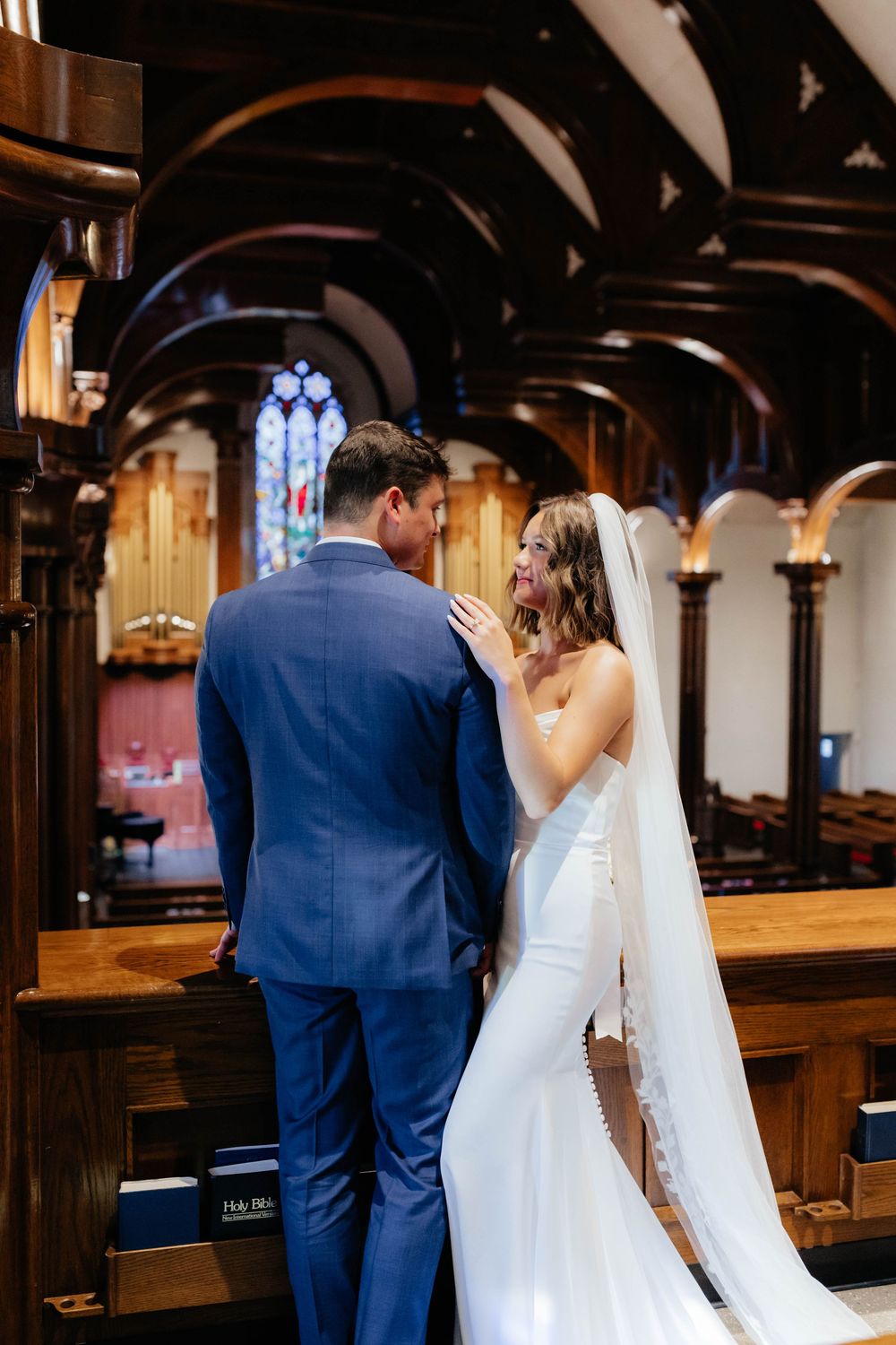 A bride and groom share an intimate moment in a church with beautiful stained glass windows and wooden pews.