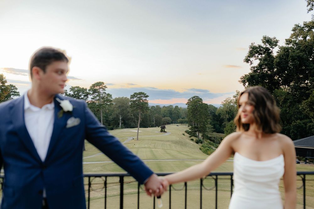 Romantic couple in wedding attire holds hands while walking through a scenic countryside landscape at sunset.