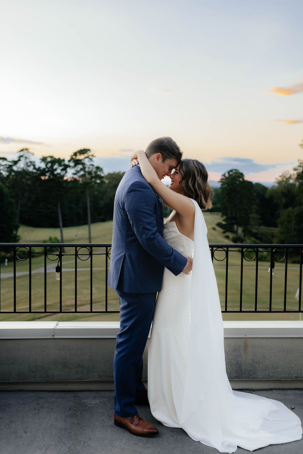 A romantic wedding couple shares an intimate moment on a balcony at sunset overlooking a scenic landscape.