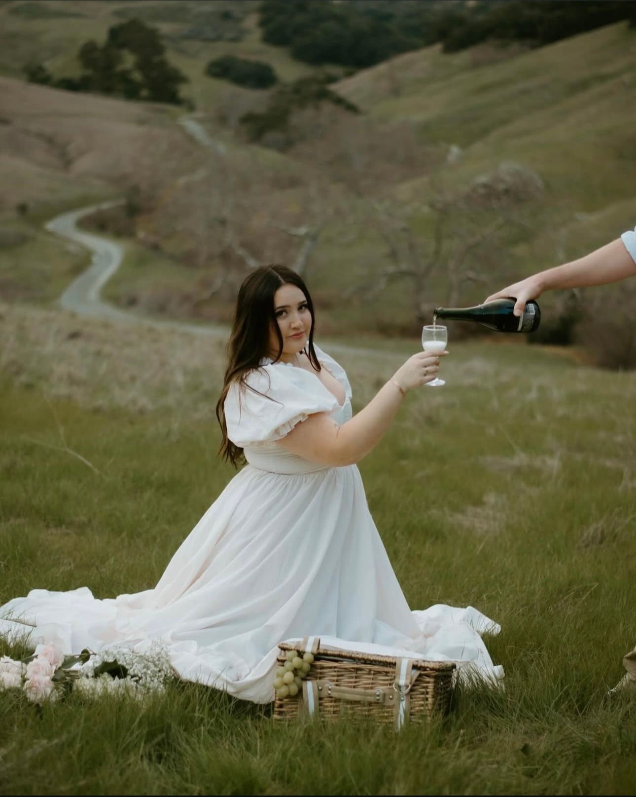 A flowing white dress twirls gracefully across a scenic green hillside landscape at sunset.