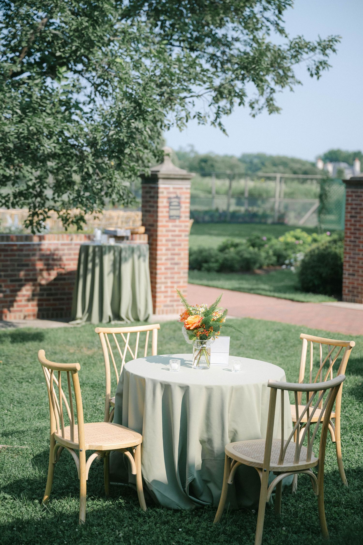 Elegant outdoor table settings with cream tablecloths and wooden chairs arranged on a green lawn near brick pillars. - Elawa Farm