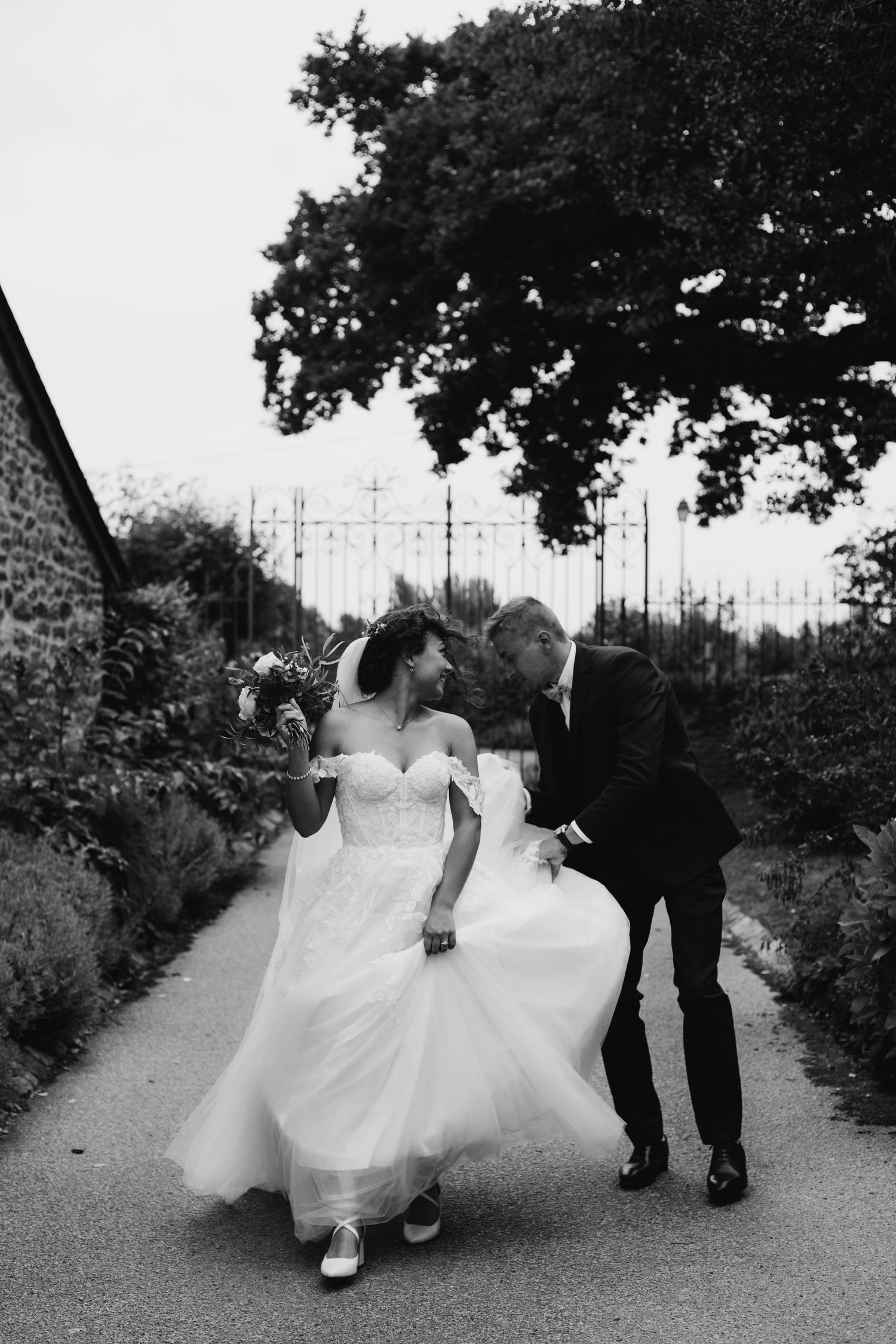 Black and white photo of bride and groom walking and laughing together on a path lined with stone walls.