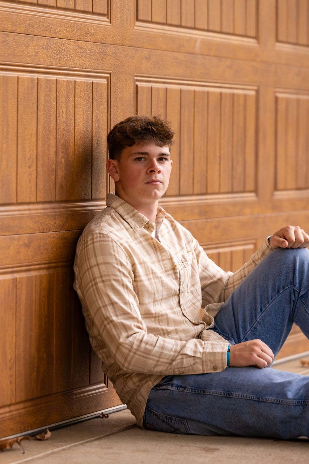 A casual portrait of someone sitting against wooden paneling wearing a plaid shirt and jeans.