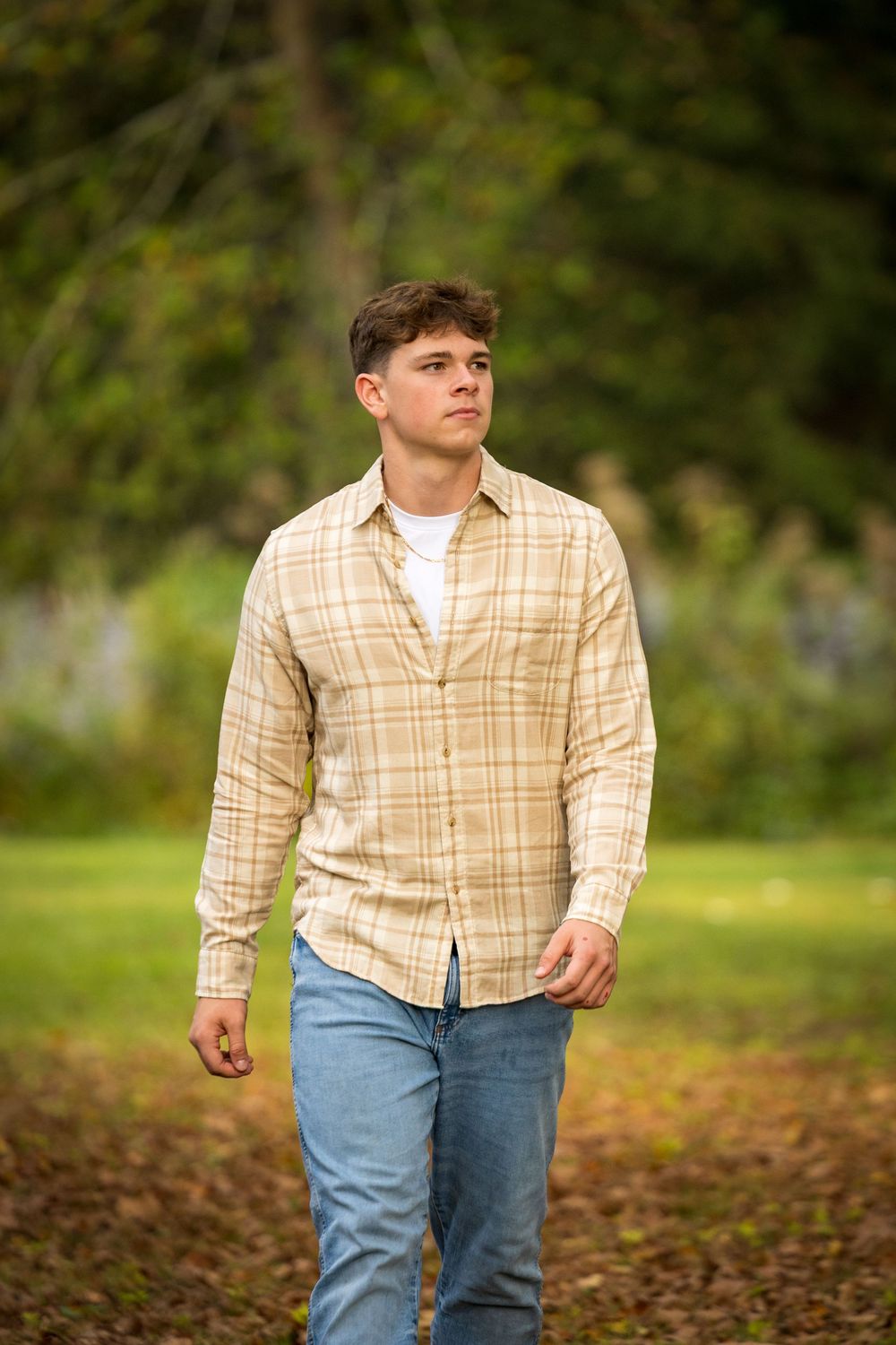 A casual outdoor portrait of someone walking on fallen leaves wearing a plaid shirt and jeans.