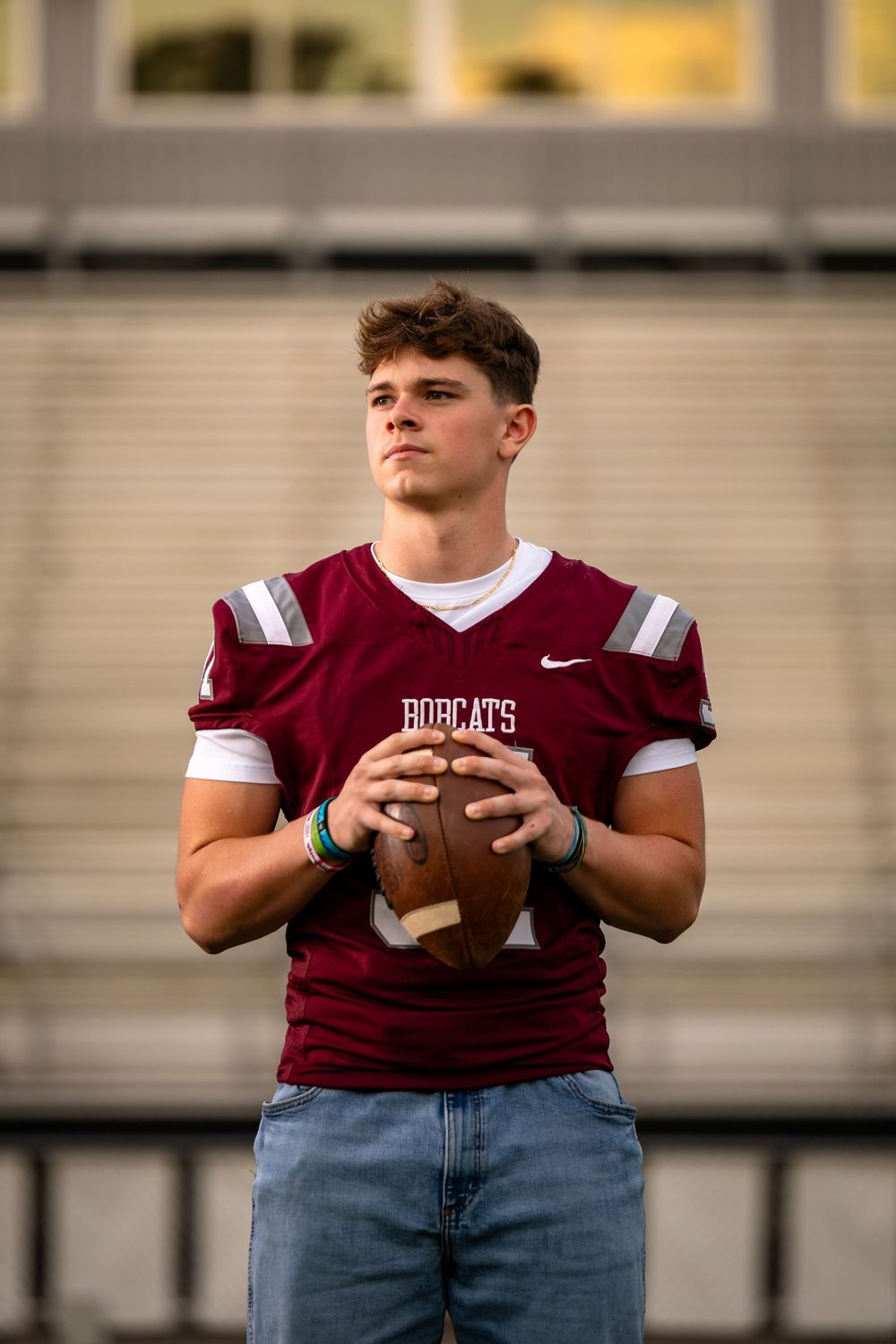High school football player in maroon jersey and jeans holds football while posing for sports portraits.
