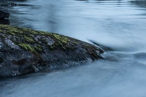 Long exposure photograph shows silky smooth flowing water around mossy rocks in a river or stream.