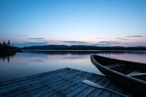 Scenic lake landscape at dusk with silhouetted boats and wooden dock against tranquil water and mountain backdrop.