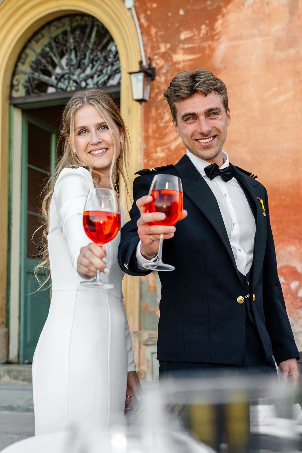 Happy couple in wedding attire toasting with red wine glasses outdoors near a colorful building.