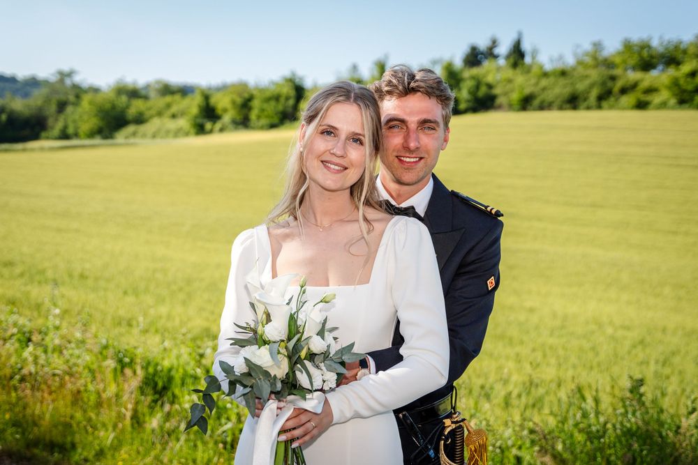 Smiling bride and groom posing in green field, bride holding white tulip bouquet, groom in kilt.