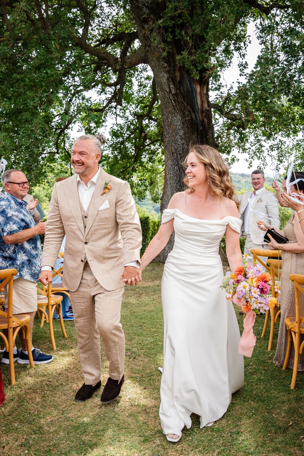 Bride and groom walk hand-in-hand down outdoor aisle under oak tree, guests celebrating at sunny garden wedding.