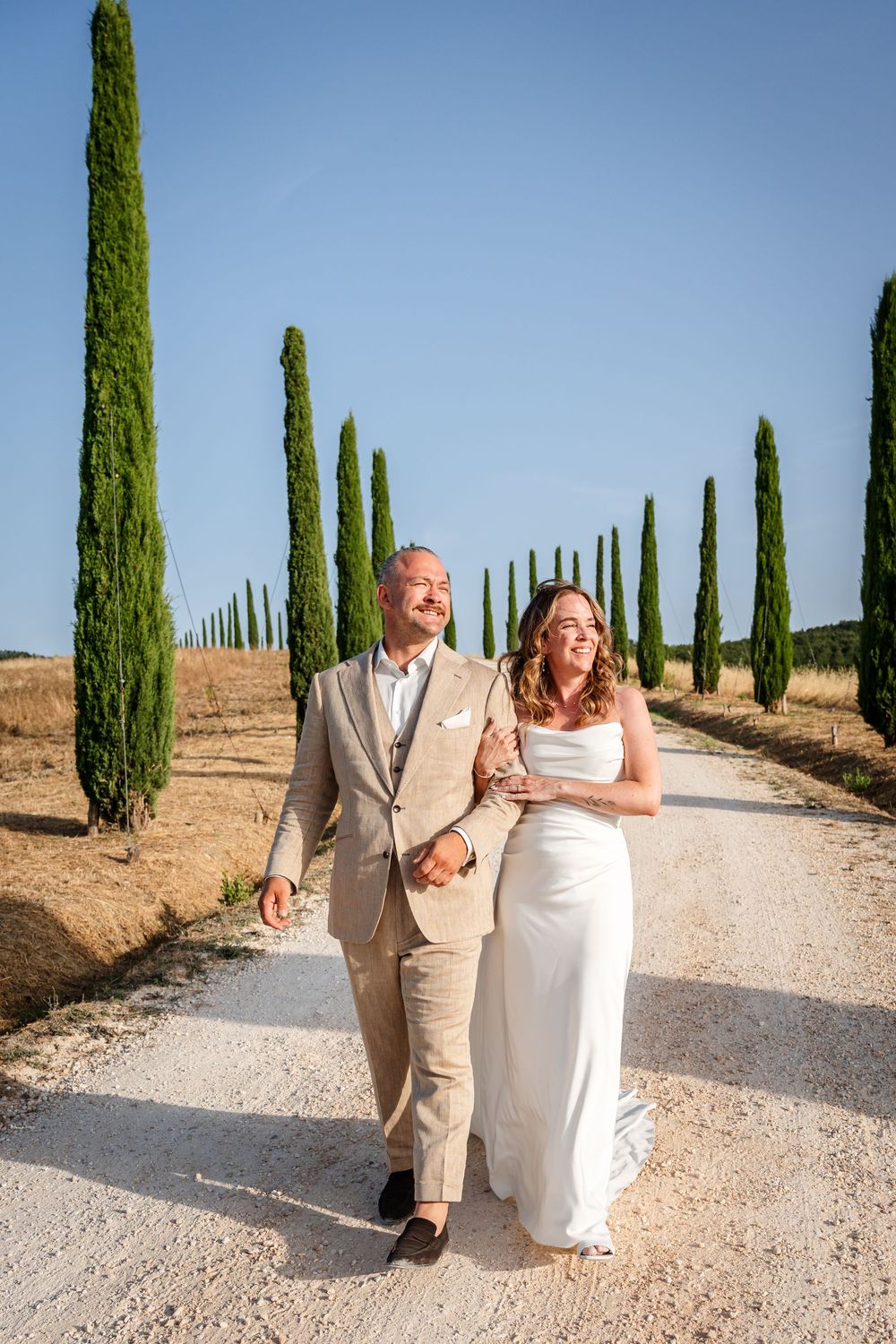 Bride and groom walking hand in hand along a cypress tree-lined path in Tuscany on their wedding day.