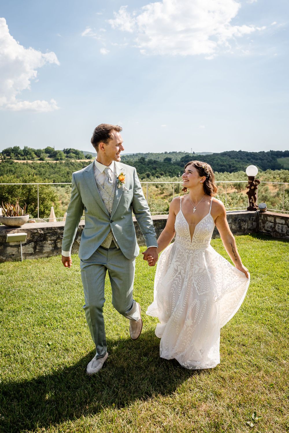 Happy newlyweds holding hands and walking on green lawn with scenic vineyard landscape behind them.