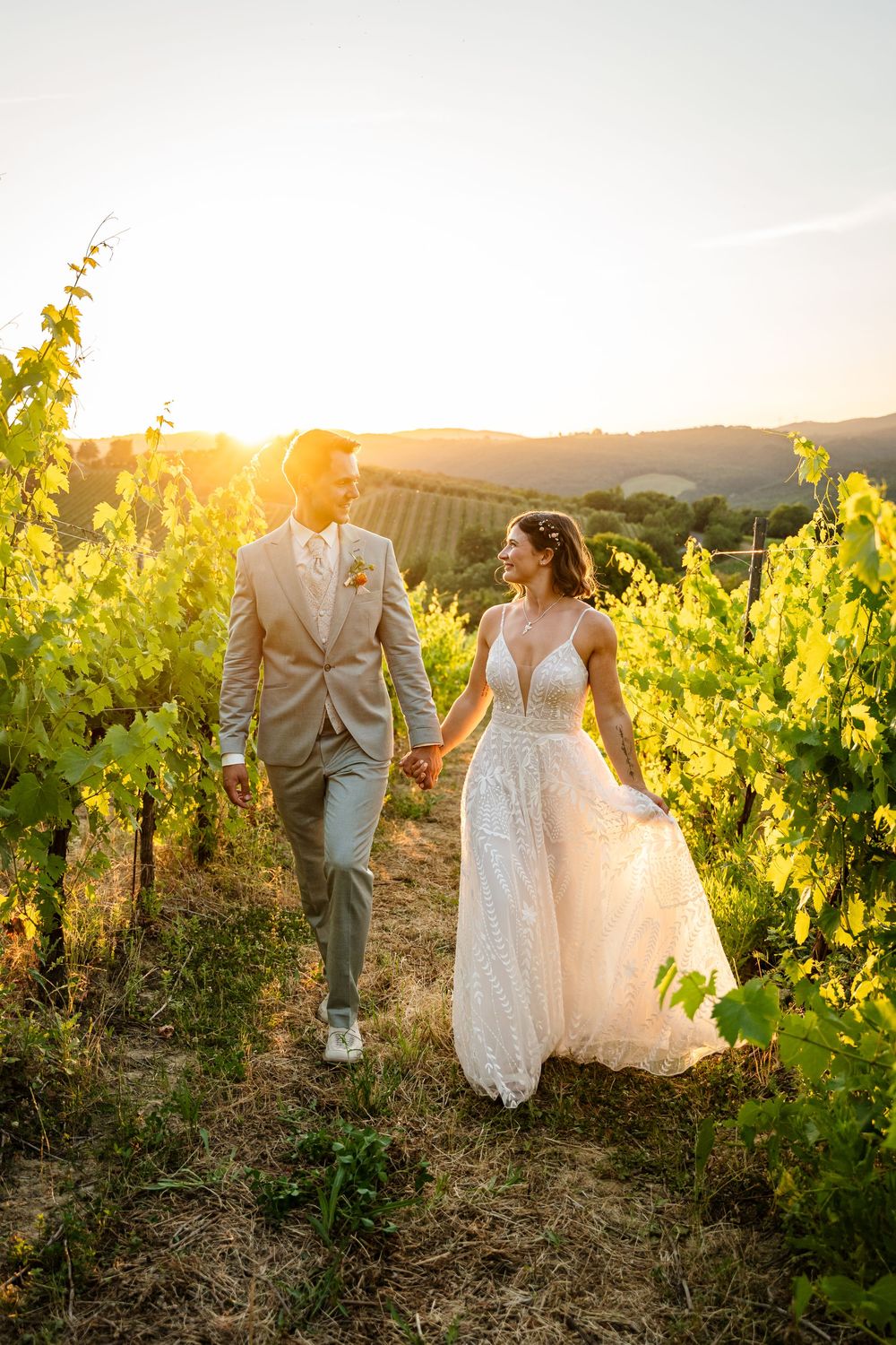 Bride and groom walk hand-in-hand through a sunlit vineyard at golden hour, bride in lace gown, groom in tan suit.