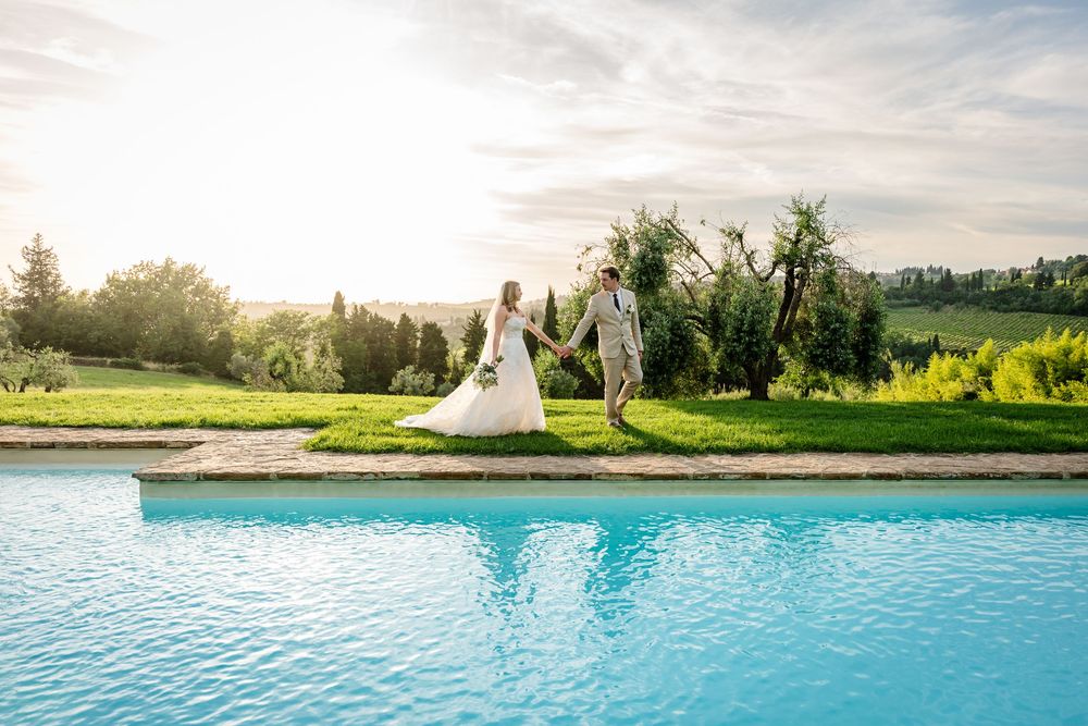 Bride and groom hold hands walking beside a turquoise pool at a sunny Tuscan villa wedding.