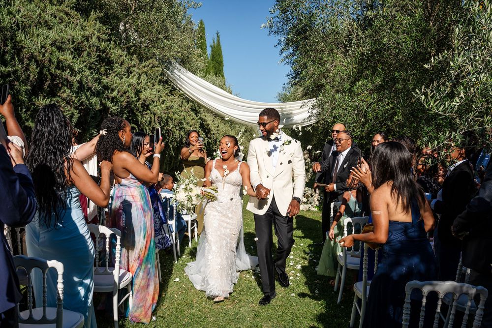 Newlyweds joyfully walking down aisle surrounded by cheering guests at outdoor garden wedding ceremony.