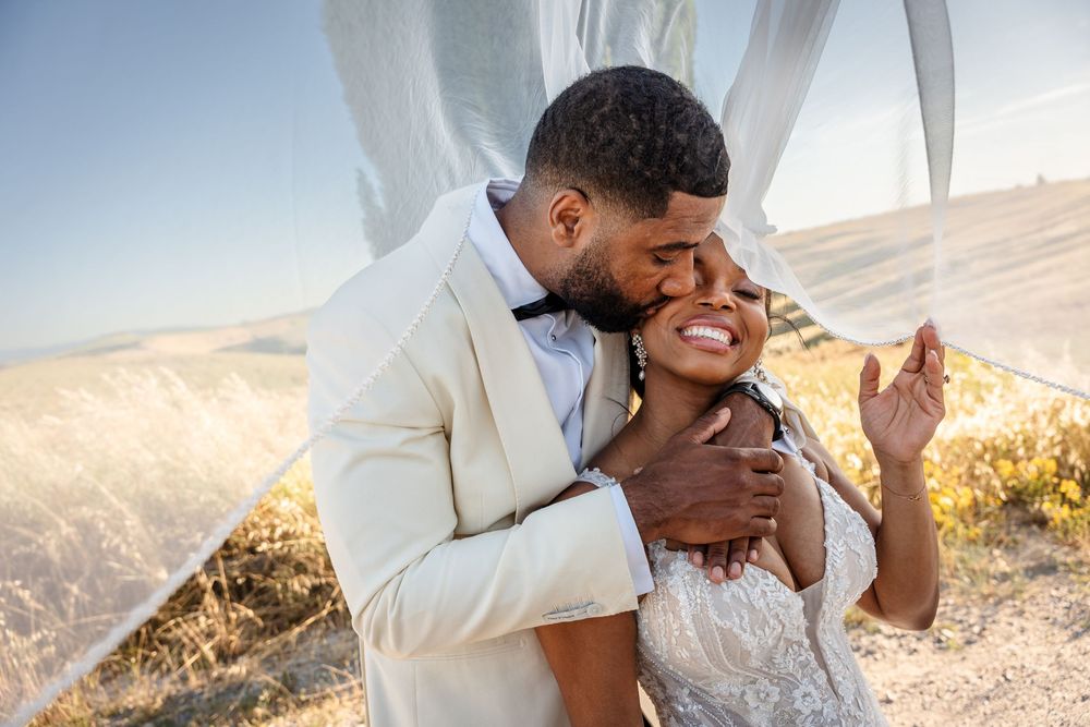 Groom in ivory suit embraces bride in lace dress under flowing white fabric in golden field.