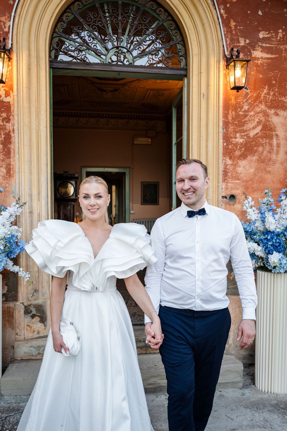 Bride in ruffled white gown and groom in navy suit hold hands outside elegant venue door with blue floral arrangements.