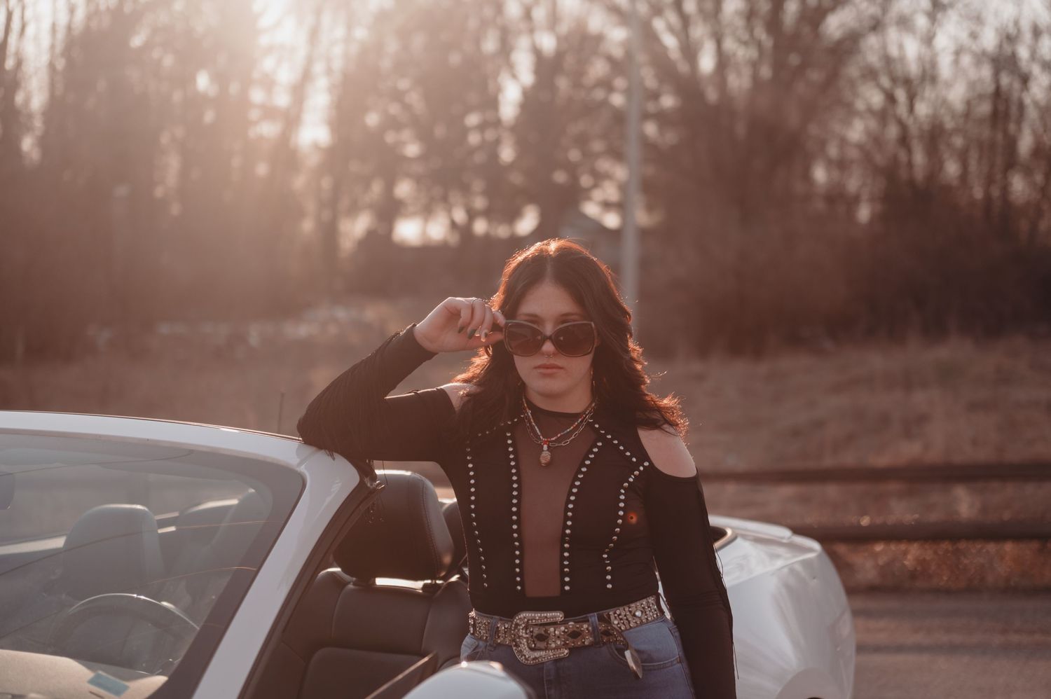 Stylish woman in sunglasses and leather jacket poses next to convertible car at sunset.