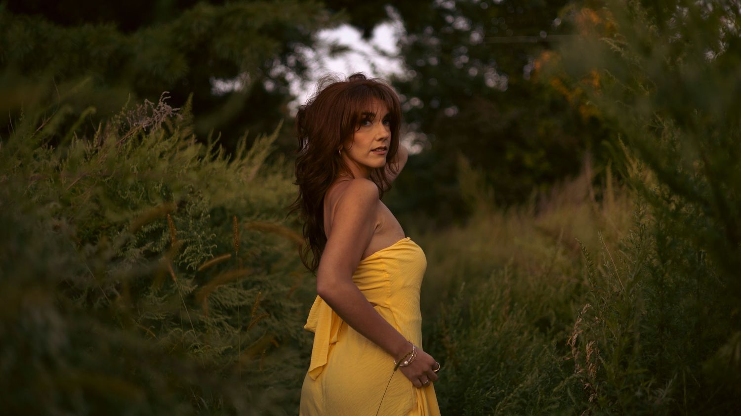 A person in a yellow dress stands in a sunlit field of tall grass during golden hour.