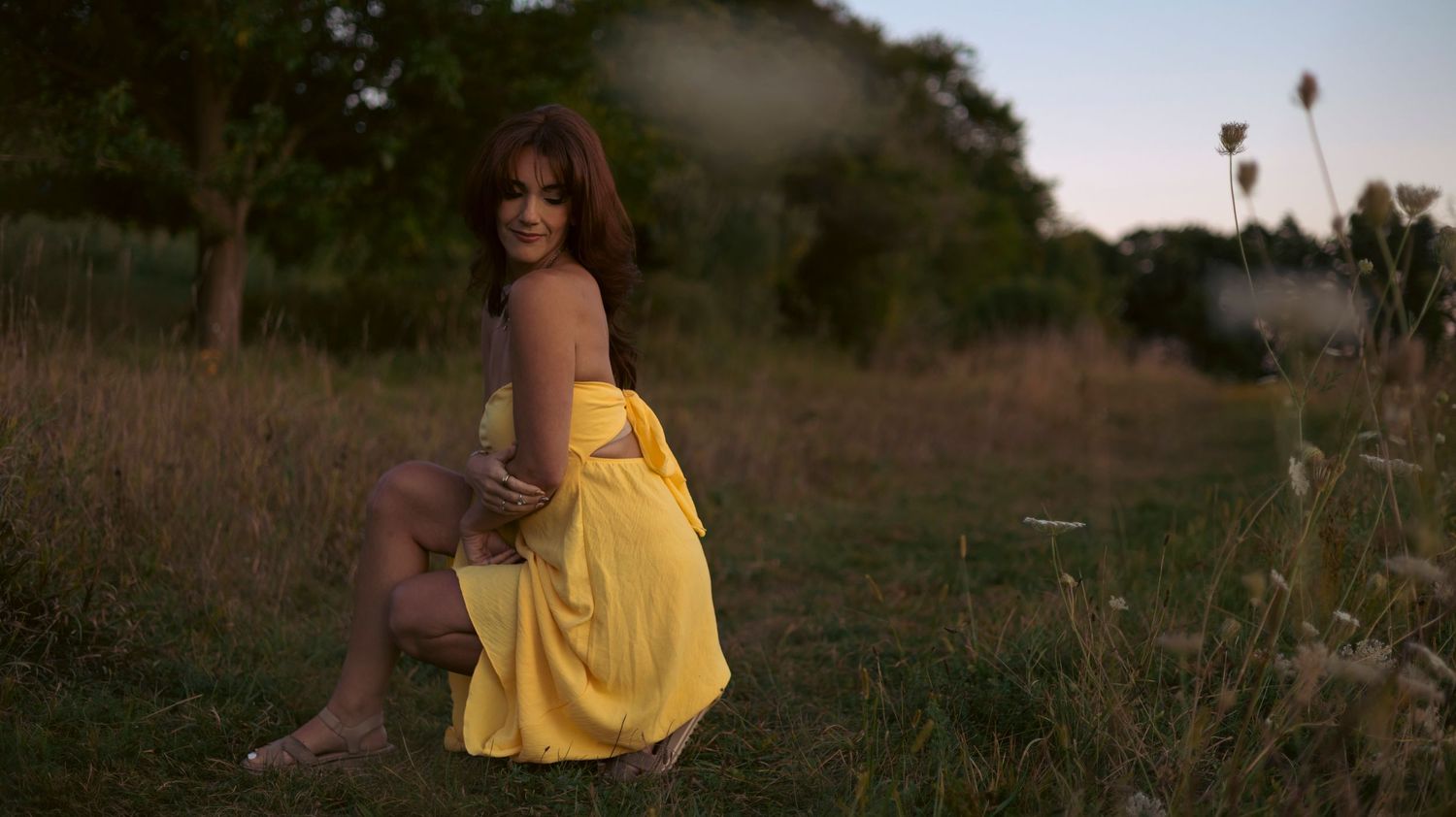 Woman in yellow dress sits in a meadow at sunset among tall grasses and wildflowers.